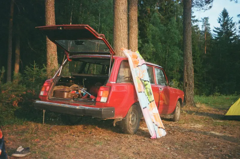 Vintage red car with open trunk in a forest, surrounded by camping gear and trees