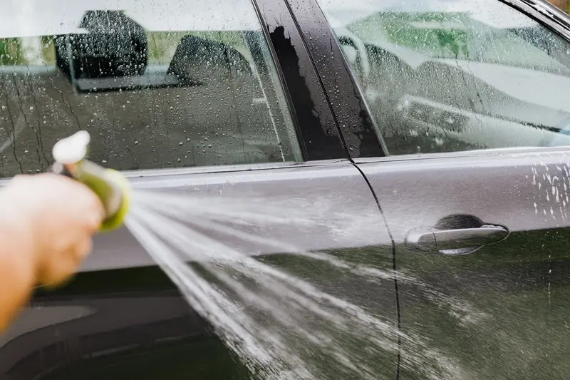 A person uses a hose to wash a black car outdoors, creating water splashes