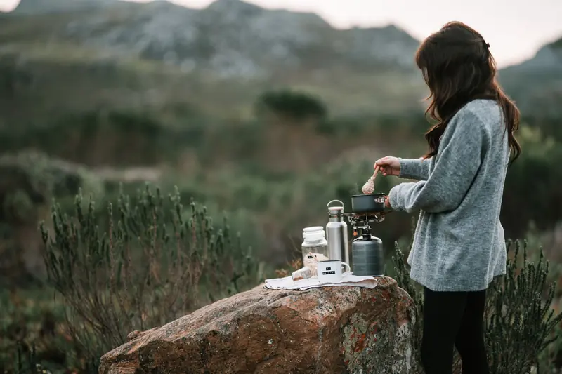 A woman prepares a warm breakfast on a portable camp stove amidst scenic outdoor surroundings