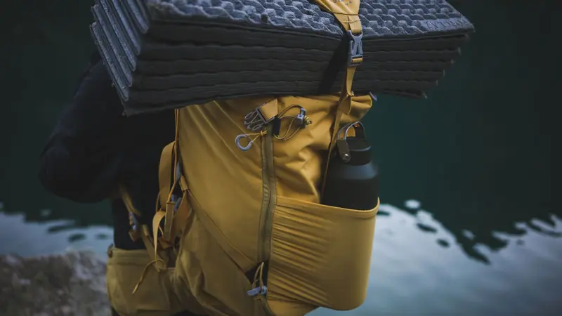 Yellow backpack with camping gear including a water bottle and folded mat near water