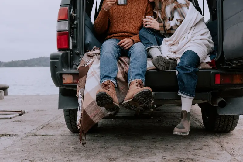 A couple enjoys a warm drink while sitting in an SUV trunk by the serene lakeside on a chilly day