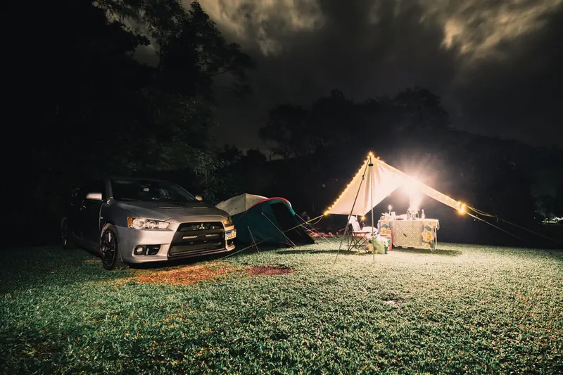 Serene camping site at night featuring illuminated tents, car, and lush grass under a cloudy sky