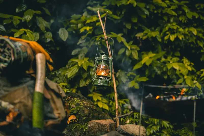 A rustic lantern hangs, casting warm light amidst lush foliage and camping gear