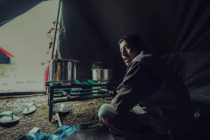 A man sits inside a tent, cooking with pots on a portable stove during a camping trip