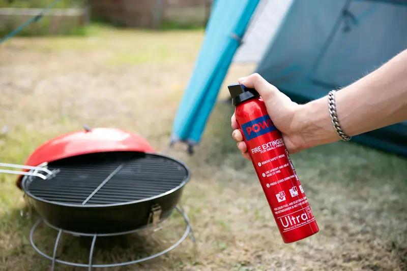 Close-up of hand holding fire extinguisher near a barbecue grill next to a tent
