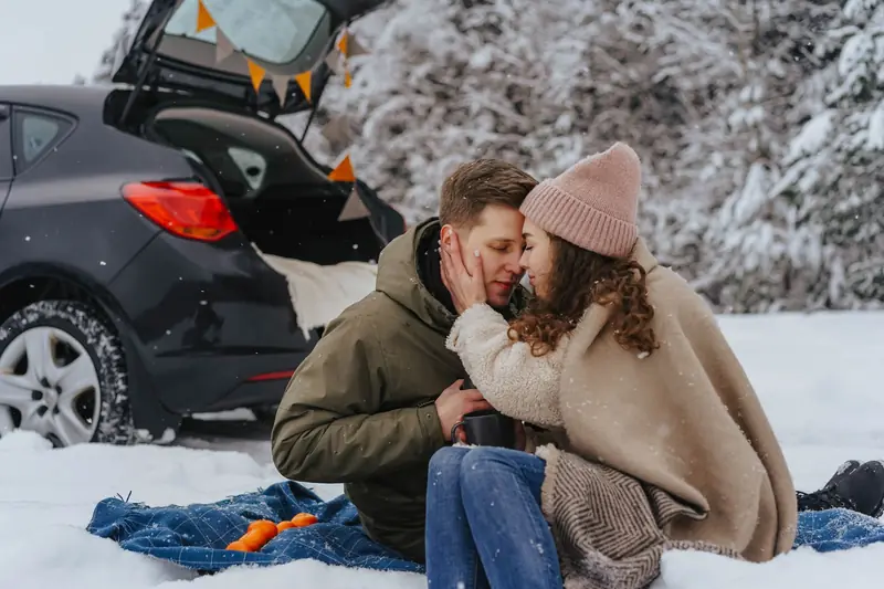 Couple enjoying a winter picnic with oranges and blanket beside open car trunk, surrounded by snow