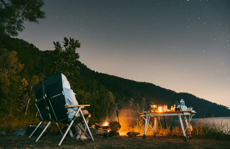 A serene camping scene by a lake with a campfire, chairs, and a star-filled sky