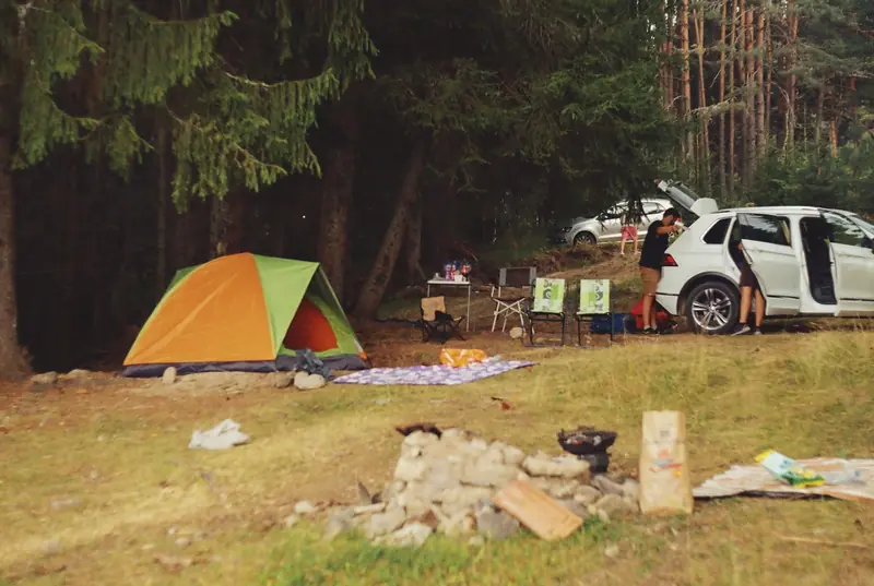 Explore a peaceful camping scene in the forest with a tent, car, and campfire setup