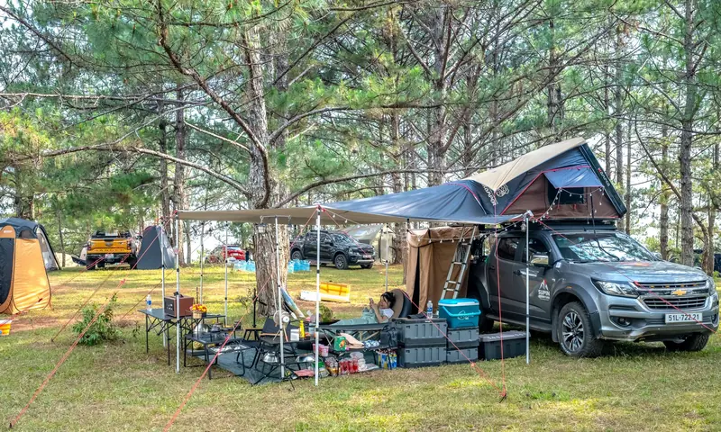 Outdoor camping scene featuring cars, tents, and a rooftop tent in a wooded area
