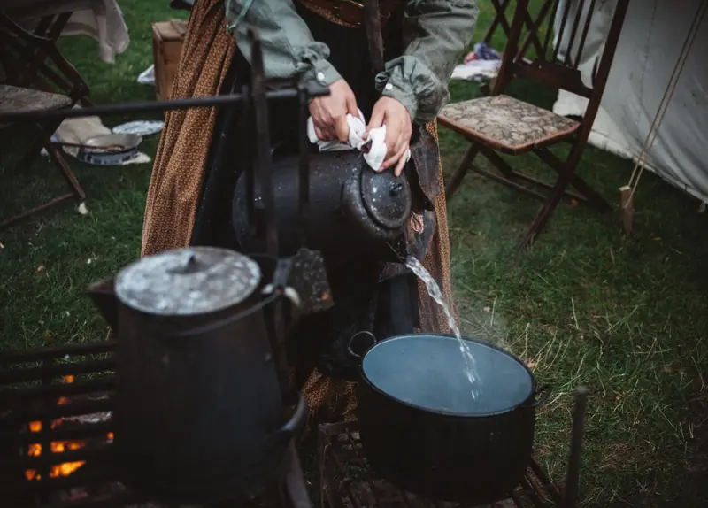 Woman pouring water from a kettle into a pot outdoors during a camping setup. Warm, rustic scene